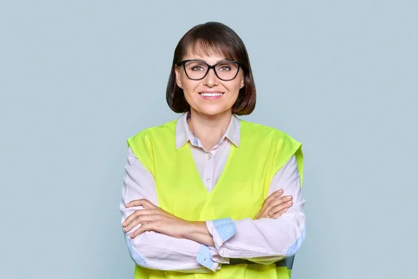 Portrait of confident industrial woman in safety vest looking at camera with crossed arms on light gray isolated background. Engineering, construction, sales, job management, work staff concept