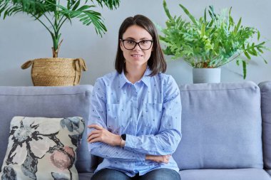 Gözlüklü genç bir kadın kanepeye oturmuş kameraya bakıyor. Kollarını kavuşturup gülümseyen kendine güvenen bir kadın, insanlara konsepti öğreten bir işletme öğrencisi.