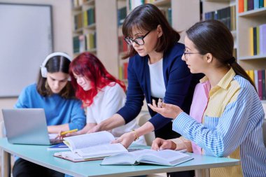 Bir grup genç öğrenci okul kütüphanesi sınıfında ders çalışıyor. Genç kız ve bayan öğretmen derslerine yardım ediyor. Lise, öğrenim, eğitim, bilgi, ergenlik
