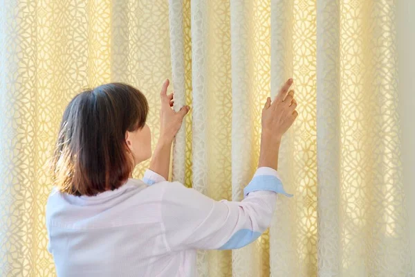 A woman touches curtains, lays out folds on light velvet curtain with lining. Window decor, curtains in home interior, natural fabrics