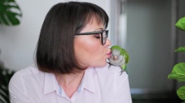 Middle aged woman at home with a pet parrot on her shoulder. Female owner talking to a bird, kissing a green Quaker parrot. Exotic birds, pets, friendship, care, love concept