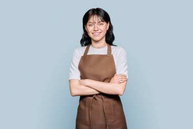 Önlüklü, kendine güveni tam genç bir kadın çalışan gri renkli arka planda kameraya bakıyor. Kollarını kavuşturmuş pozitif kadın işçi, öğrenciler ve kiralık iş, servis personeli konsepti