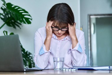 Tired middle-aged woman suffering from headache, overwork, sitting at workplace with laptop. Mature female stressed, migraine, overworked female supervisor, worker working remotely in home office