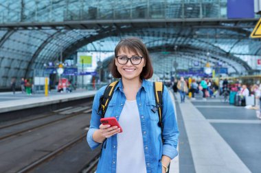 Woman passenger with smartphone waiting on railway platform inside station, smiling female with backpack looking at camera. Rail transport, passenger transportation, journey tourism travel trip people