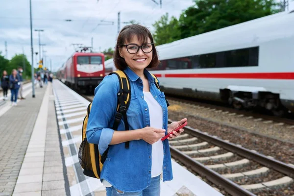 Woman passenger waiting for commuter train at station outdoor platform, smiling female looking at camera. Rail transport, passenger transportation, journey, tourism, travel, trip, people concept