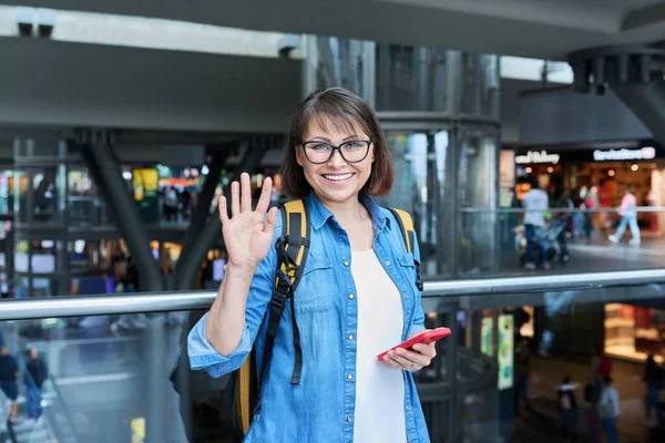 Modern istasyon binasında sırt çantalı akıllı telefonlu orta yaşlı bir kadın. Şehir mimarisi, şehir yaşam tarzı, ulaşım, yolcu taşımacılığı, gidiş yeri seyahat insanları kavramı