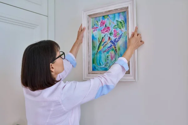 Woman hanging floral birds art framed at home on the wall. Art, decoration, interior design, paintings in the cozy house