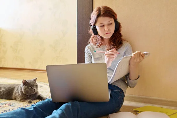 Preteen girl studying at home using laptop along with cat. Child in headphones sits on floor with notebooks, pet plays lies with young owner. Animals, friendship, children, lifestyle, study, school