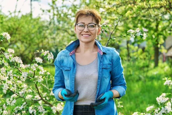 Portrait of beautiful middle aged woman in blooming spring garden. Smiling female in gardening gloves looking at camera. Springtime, beauty, nature, orchard, 40s people concept