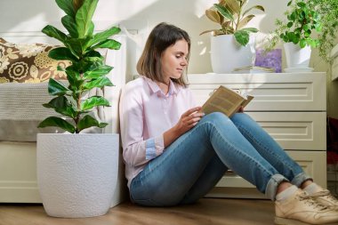 Evde yerde oturmuş kitap okuyan genç bir kadın. Hevesli, gevşemiş kadın kitap okumaktan zevk alıyor, evde, hafta sonu, hobi, boş zaman, eğitim kavramı.