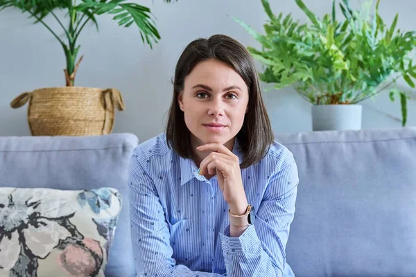 Young beautiful woman looking at camera while sitting on couch at home. Portrait of smiling business female, teacher worker freelancer student blogger vlogger. Beauty, work, job, people 20s concept
