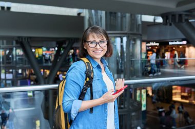 Modern istasyon binasında sırt çantalı akıllı telefonlu orta yaşlı bir kadın. Şehir mimarisi, şehir yaşam tarzı, ulaşım, yolcu taşımacılığı, gidiş yeri seyahat insanları kavramı