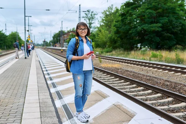 Woman passenger waiting for commuter train at station outdoor platform, smiling female looking at camera. Rail transport, passenger transportation, journey, tourism, travel, trip, people concept