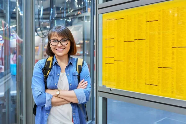 Female passenger with a backpack looking at the camera standing near the board with the timetable of transport, suburban trains at the railway station