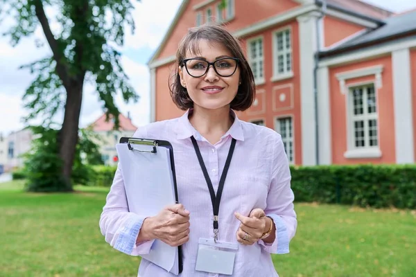 Middle-aged positive female school worker, teacher on background of school building. Woman psychologist advisor social worker with fileholder looking at camera outdoor. Education staff people concept