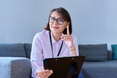 Ofisteki koltukta oturan kadın psikoloğun portresi. Kadın terapist, elinde not panosuyla kameraya gülümseyen sosyal hizmet görevlisi. Profesyonel yardım desteği, psikoloji, psikiyatri, ruh sağlığı