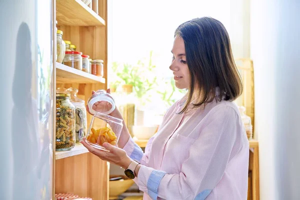 Young woman in kitchen with dried apricots, containers jars of cereals, paste, spice, dry fruits. Female in home pantry, food organization and storage, kitchen utensils, household concept