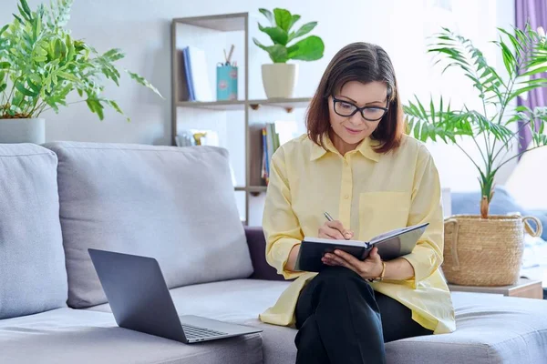 Mature businesswoman working on couch, using laptop making notes in business notebook. Confident middle aged female working from home office remotely. Business job freelancing, technology, 40s people