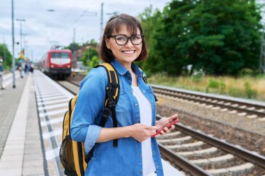 Woman passenger waiting for local train at station outdoor platform, smiling female looking at camera. Rail transport, passenger transportation, journey, tourism, travel, trip, people concept