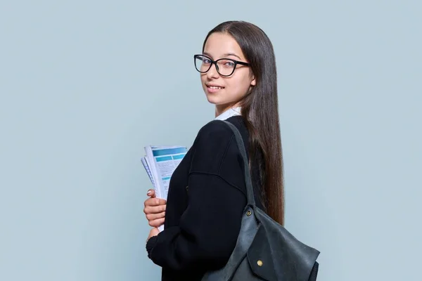 Portrait of teenage female student with backpack notebooks looking at the camera on gray color background. Attractive teen girl in glasses posing in studio. High school, adolescence, study, education