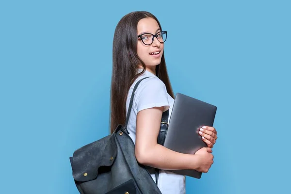 Portrait of teenage female student with backpack laptop looking at camera on blue color background. Attractive teen girl in glasses posing in studio. High school, adolescence, study, education