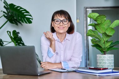 Orta yaşlı, gülümseyen bir iş kadınının portresi masada dizüstü bilgisayarla oturuyor. Gözlüklü güzel olgun bir kadın kameraya bakıyor, iş yerinde çalışıyor, serbest çalışıyor, 40 'lı yaşlarda insanlar konsepti var.