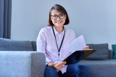 Ofisteki koltukta oturan kadın psikoloğun portresi. Kadın terapist, elinde not panosuyla kameraya gülümseyen sosyal hizmet görevlisi. Profesyonel yardım desteği, psikoloji, psikiyatri, ruh sağlığı