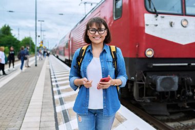 İstasyonda tren bekleyen bir kadın yolcu, kameraya gülümseyen bir kadın. Demiryolu taşımacılığı, yolcu taşımacılığı, yolculuk, turizm, seyahat, seyahat, insan konsepti