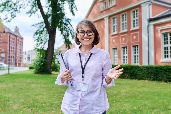 Middle-aged positive female school worker, teacher on background of school building. Woman psychologist advisor social worker with clipboard looking at camera outdoor. Education staff people concept