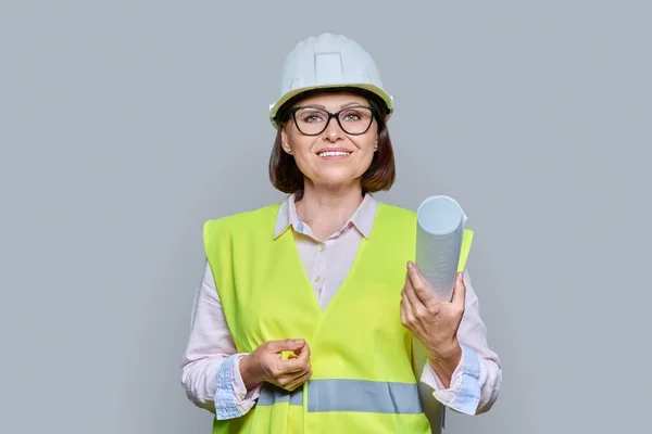 Portrait of female industrial worker with tech blueprint, wearing safety vest and hard hat, on gray color background. Construction, engineering, management, building, development, staff, industry