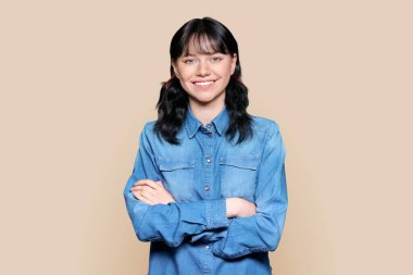 Portrait of beautiful teenage student woman looking at camera. Young charming brunette female smiling with teeth, confident with arms crossed on light brown color isolated background