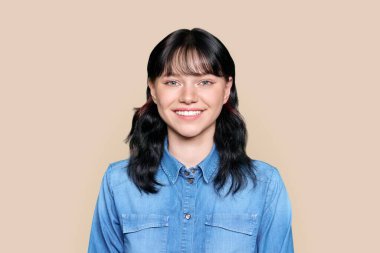 Joyful happy young teenage girl looking at camera on light brown color isolated background. Beautiful female brunette 17, 18 years old, smiling with teeth, in denim shirt. Youth, joy, success, beauty