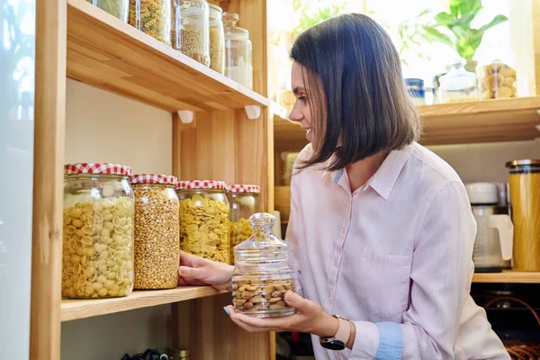 Young woman in kitchen with containers jars of nuts, cereals, pasta, dry fruits. Female in home pantry, food organization and storage, kitchen utensils, household, food preparation at home concept