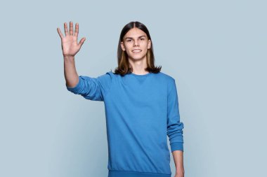 Young guy looking at camera waving his hand, on grey color background. Handsome student teenage guy posing in studio. Youth, greeting hello hi, people concept