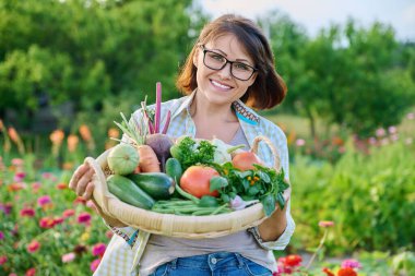 Portrait of smiling middle aged woman with basket of different fresh raw vegetables and herbs, summer nature vegetable garden background. Harvest from organic farm, healthy food nutrition, gardening