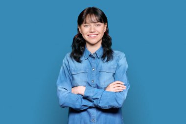 Portrait of beautiful teenage student woman looking at camera. Young charming brunette female smiling with teeth, confident with arms crossed on light blue color isolated background