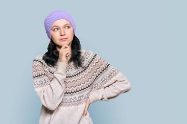 Portrait of pensive young female touching her chin with hand, in winter sweater hat, looking away creating solution copy space, on gray color isolated background. Question doubt reflection choice