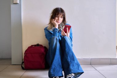 Sırt çantalı, akıllı telefonlu üzgün kız öğrenci asansörün yanında oturuyor. Yalnız, hayal kırıklığına uğramış genç bir kadın, hayal kırıklığı, acı depresyonu konsepti.