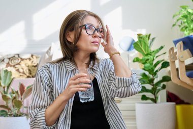 Evde bir bardak suyla baş ağrısı çeken olgun bir kadın. Sağlık, stres, depresyon, acı, migren, yaşlılık sorunları