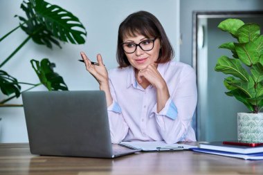 Video görüşmesi için dizüstü bilgisayar kullanan olgun bir kadın, sanal toplantı, konferans sohbeti, bilgisayar ekranından konuşma. Orta yaşlı, gözlüklü iş kadını ofis masasında oturuyor.