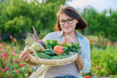 Portrait of smiling middle aged woman with basket of different fresh raw vegetables and herbs, summer nature vegetable garden background. Harvest from organic farm, healthy food nutrition, gardening