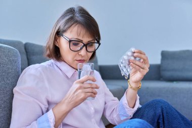Olgun hasta bir kadın bir bardak suyla ilaç alıyor. Ağrı kesiciler, ağrı kesiciler, vitaminler, antibiyotikler, antidepresanlar, acil tedavi ilaçları