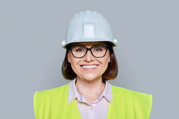 Headshot portrait of female industrial worker in protective hard hat and vest on gray color background. Construction, engineering, management, building, development, staff, industry concept