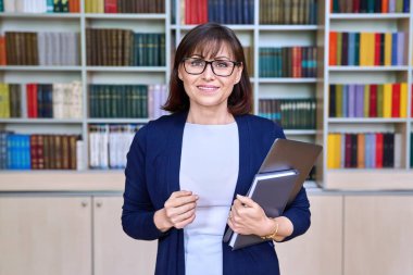 Portrait of female librarian with laptop in hands in library. Smiling middle aged female teacher in glasses looking at camera, professor mentor tutor posing in class office