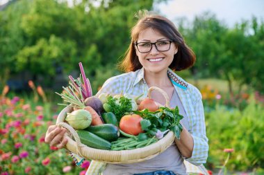 Portrait of smiling middle aged woman with basket of different fresh raw vegetables and herbs, summer nature vegetable garden background. Harvest from organic farm, healthy food nutrition, gardening