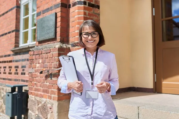 Portrait of female teacher, school worker looking at camera outdoor on steps of school building. Smiling positive woman counselor, school psychologist with clipboard. Education staff people concept