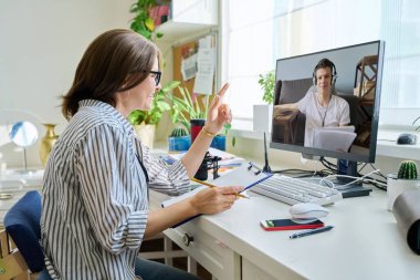 Mature woman talking online with teenage male using video call on computer, home interior. Virtual meeting, therapy session with psychologist. Chat conference technology psychology education learning