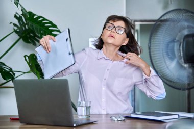 Middle-aged woman suffering from heat, overheated, cooled by fan and fan. First symptoms of menopause, age-related changes, health problems, use of vitamins, drugs, hormones to relieve