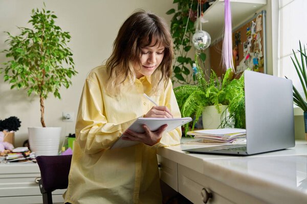 Teenage female student studying at home using laptop. Smiling girl sitting at table looking at screen writing in notebook. Education, learning, high school, college concept