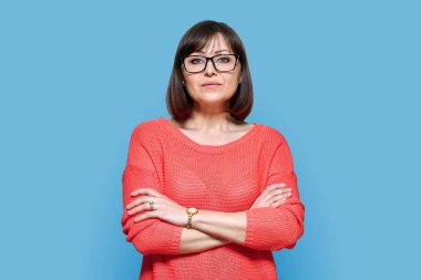 Serious confident middle aged woman in glasses on blue color background. Experienced mature female in red casual clothes looking at camera. Lifestyle, confidence, calmness, mature people concept